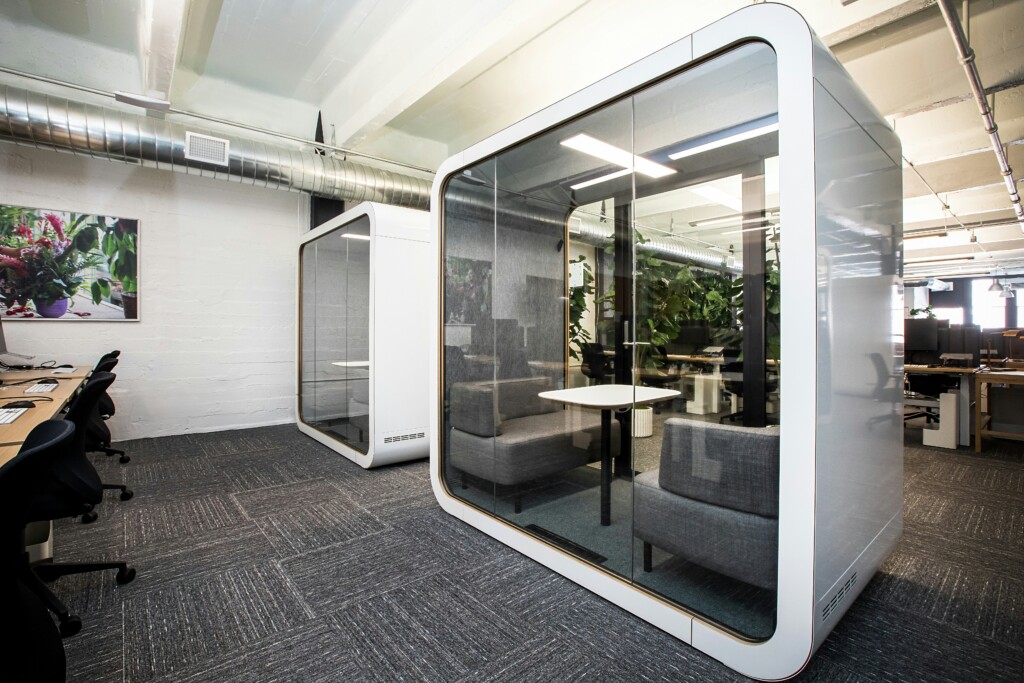 Modern office with glass meeting pods, gray seating, desks, plants and exposed industrial ceiling.