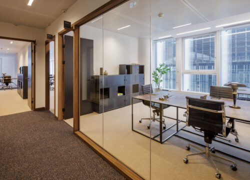 Modern glass-walled office room with dark metal desks, ergonomic chairs, modular storage, and large windows overlooking an urban exterior.