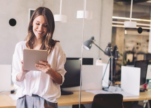 Smiling professional woman holding a tablet in a bright, modern open-plan office workspace.