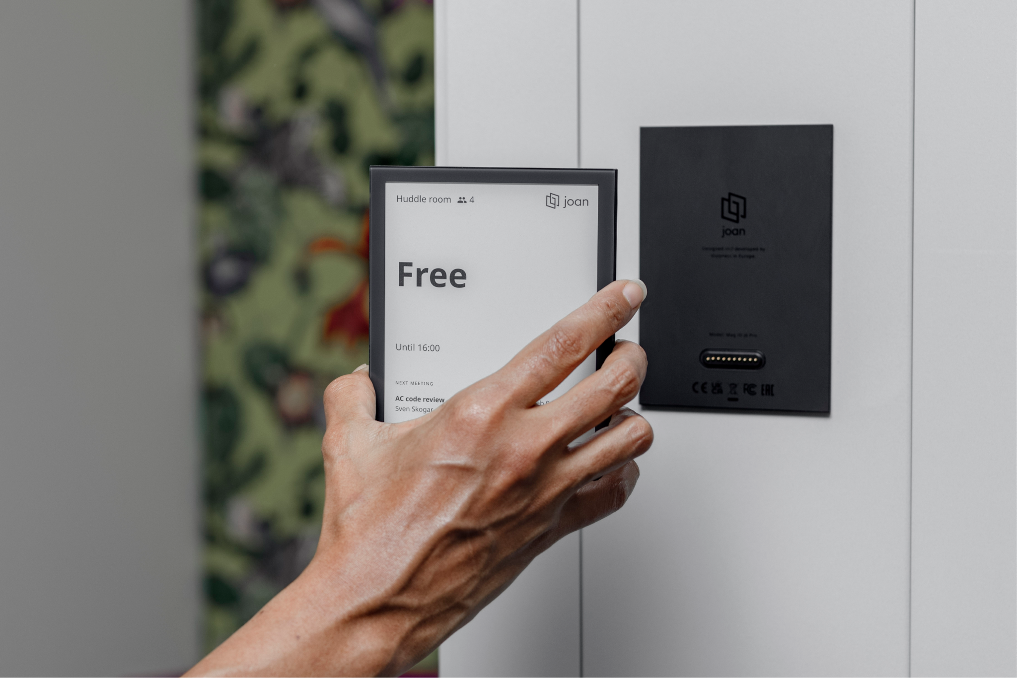Hand removing a Joan 6 Pro e-ink display from its wall mount, showing a 'Free' room status on screen.