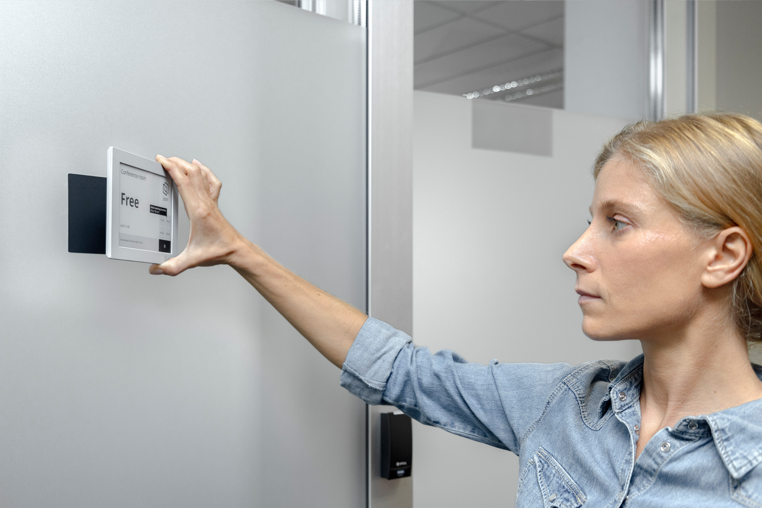 Woman interacting with a Joan 6 RE e-ink display mounted on a wall, showing a conference room is free.