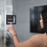 A woman interacts with a Joan 6 e-ink display showing a booked conference room schedule.
