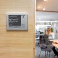 Joan 6 e-ink display showing a meeting room schedule on a wood-paneled wall next to a glass door.
