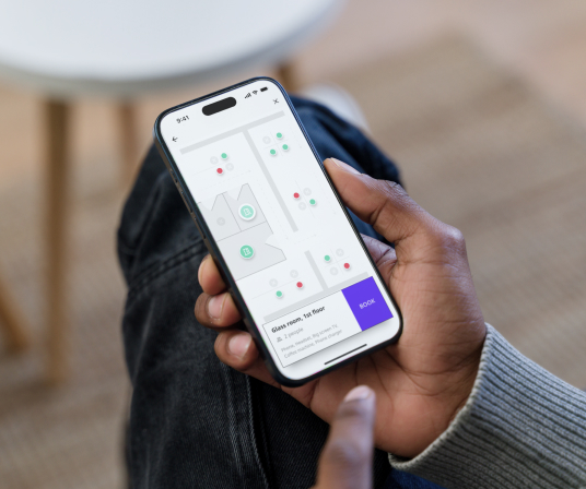 Hand holding a smartphone showing a Joan workplace management app with room or desk availability indicators.