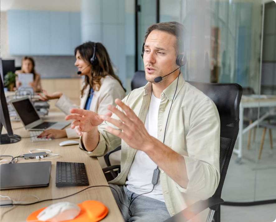 Customer support agent with headset gesturing while speaking, colleagues working in background at modern help center.